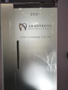 A glass door with frosted glass panels featuring vinyl graphics. The graphics include the year 2001, the Armstrong Dental Studio logo, and the name and credentials of Dr. Omar Armstrong, DDS, MS. The vinyl graphics provide a professional and informative look for the dental studio's entrance.