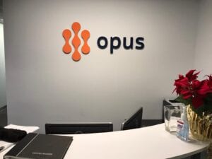 A modern office reception area featuring a wall-mounted sign for Opus. The sign includes the company logo, which is a stylized orange shape, and the company name in raised, blue letters. A white reception desk with a curved countertop is positioned in front of the wall. There are potted plants, a computer, and office supplies on the desk.