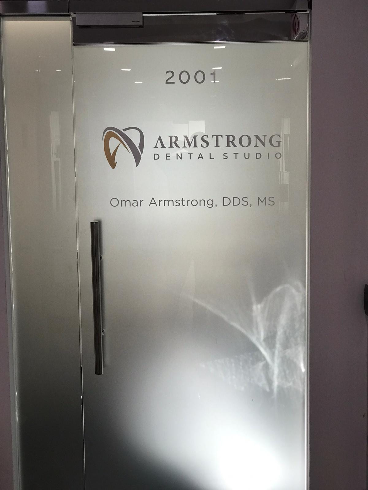 A glass door with frosted glass panels featuring vinyl graphics. The graphics include the year 2001, the Armstrong Dental Studio logo, and the name and credentials of Dr. Omar Armstrong, DDS, MS. The vinyl graphics provide a professional and informative look for the dental studio's entrance.