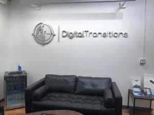 A modern office reception area featuring a wall-mounted sign for Digital Transitions. The sign includes a circular logo and the company name in raised, metallic letters. A black leather sofa, a small table, and a water cooler are also visible in the background.