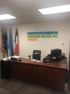 A modern office reception area with a wall-mounted sign for the Hudson River Park Trust. The sign features the organization's name in green letters with a blue underline. The American flag, the New York State flag, and the New York City flag are displayed on flagpoles behind the sign. A wooden desk, office chairs, and computers are also visible in the background.